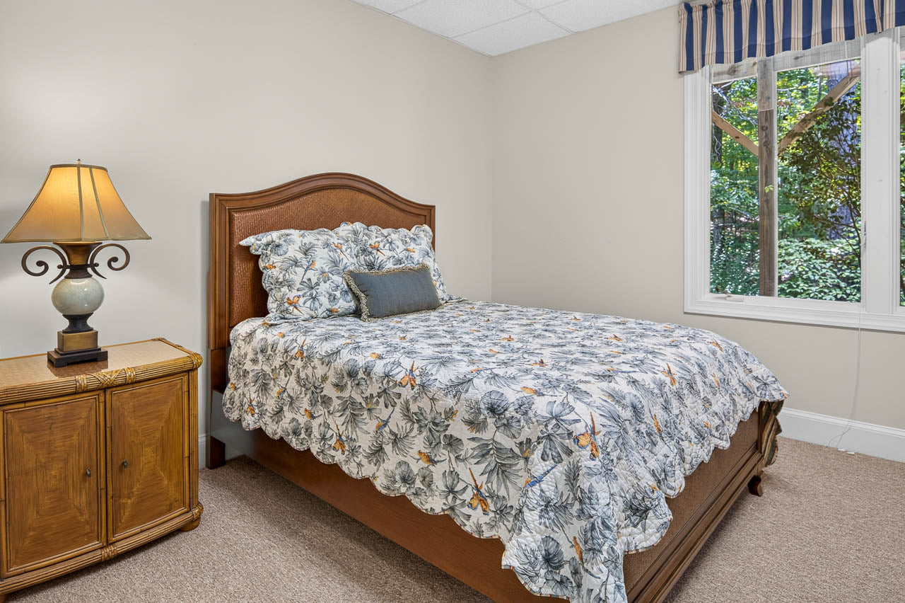 A cozy bedroom with a wooden bed, floral bedding, a decorative pillow, a lamp on a wicker nightstand, and a window with a view of green foliage outside.