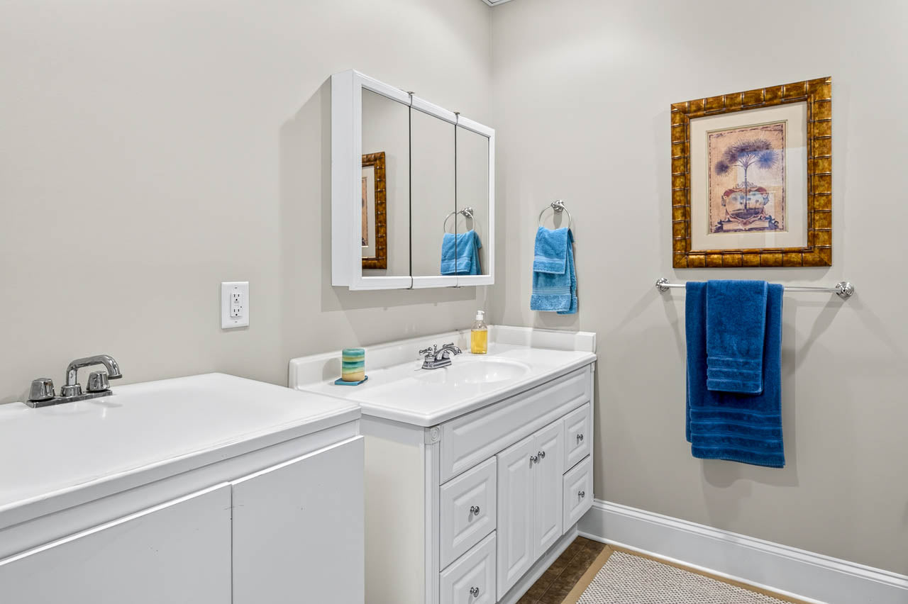 A clean bathroom with a white vanity, sink, and mirror cabinet. Blue towels hang on a rack and ring, and a framed picture is on the wall. Soap, a toothbrush holder, and a rug complete the space.