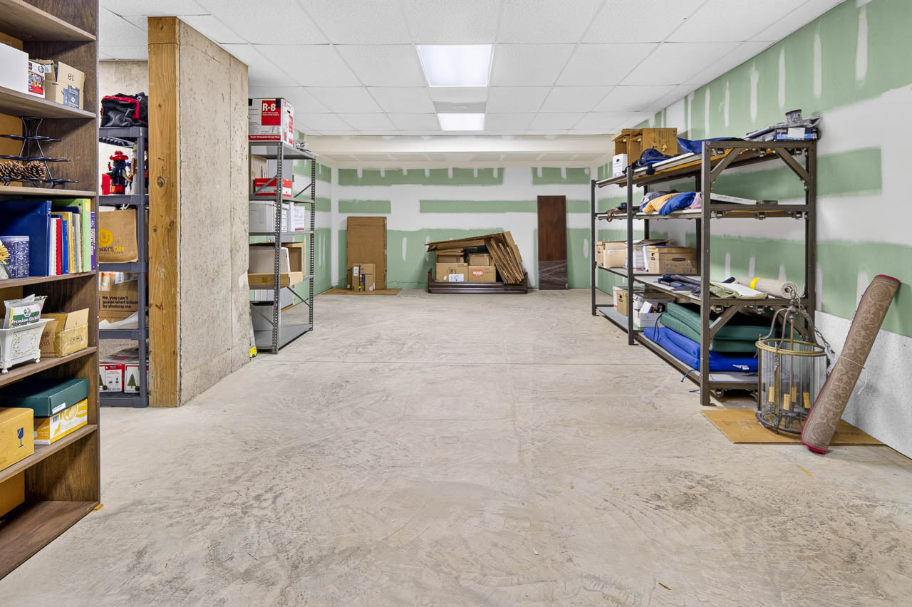 Unfinished storage room with concrete floor, green-and-white drywall, shelves of boxes and supplies along the walls, and stacked cardboard in the back. Ceiling has fluorescent lights. Room appears organized but unfinished.