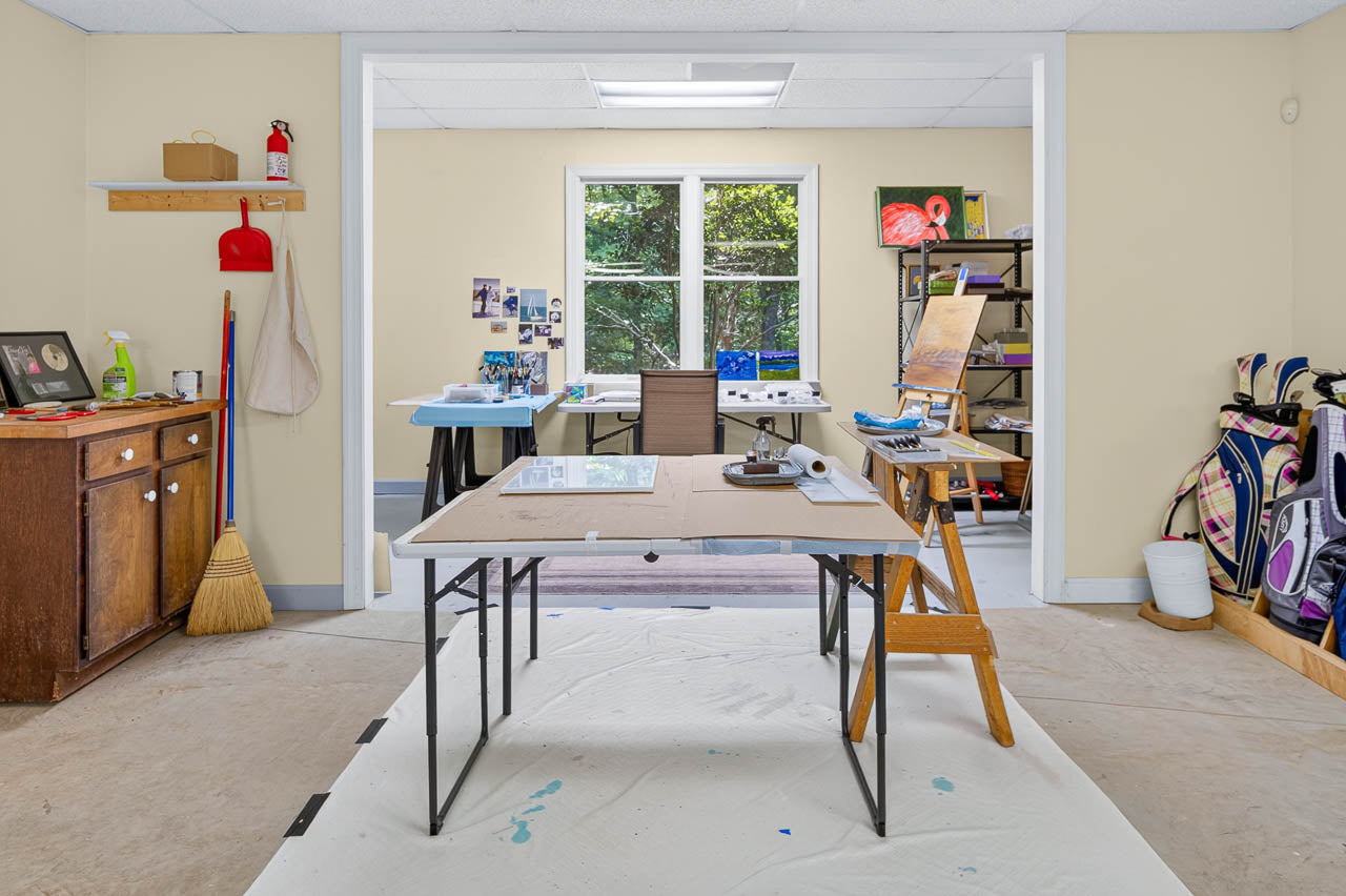 Art studio with a central work table, easel, art supplies, broom, and golf bags. A large window lets in natural light; shelves and cabinets are organized along the walls.