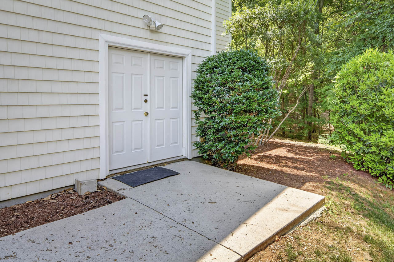 A white exterior door with three panels and a black doormat, set against light siding. Bushes and trees surround the concrete sidewalk leading to the door on a sunny day.