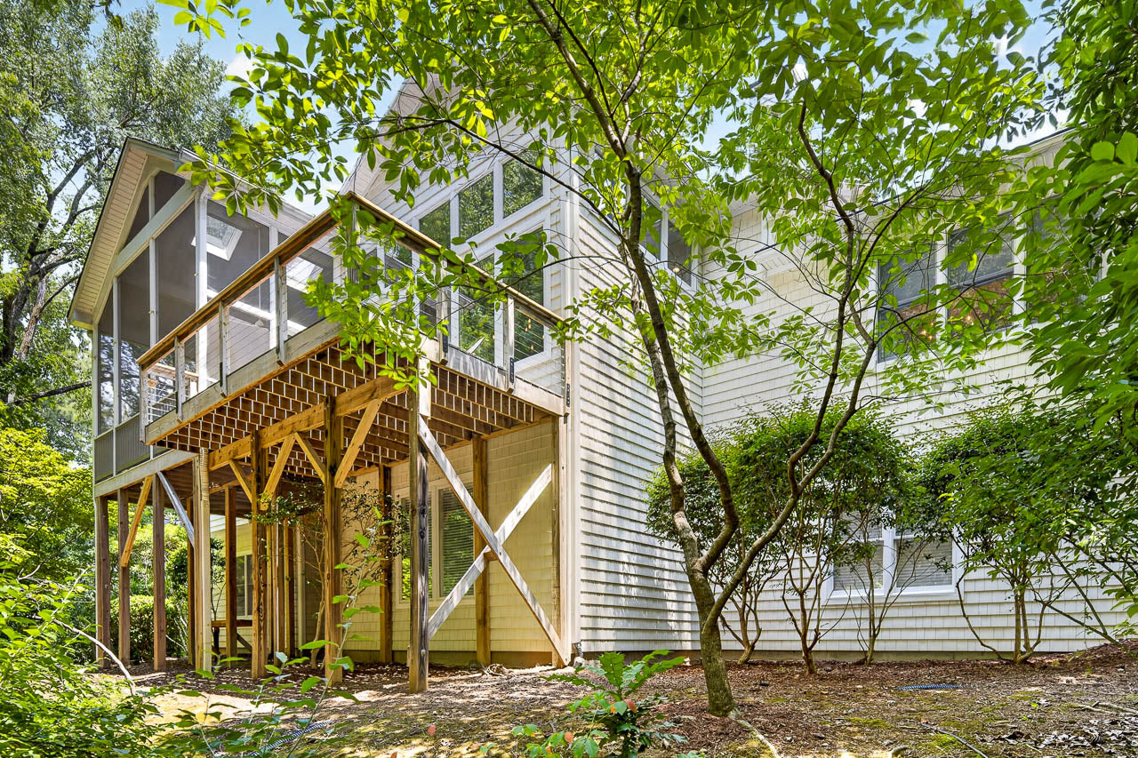 A two-story house with light-colored siding, elevated screened porch, and wooden support beams, surrounded by greenery and trees, viewed from the backyard.