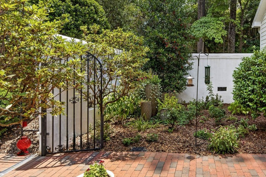 A garden area with various green plants and shrubs, a brick pathway, a black metal gate, white fence, bird feeder, and trees in the background on a sunny day.