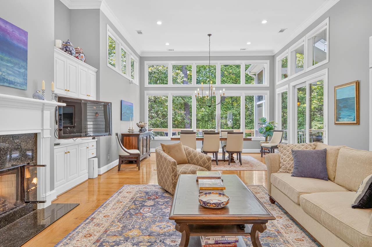 Bright living and dining room with large windows, beige sofa, patterned rug, fireplace, wall art, and a view of greenery outside; elegant, airy, and modern decor.