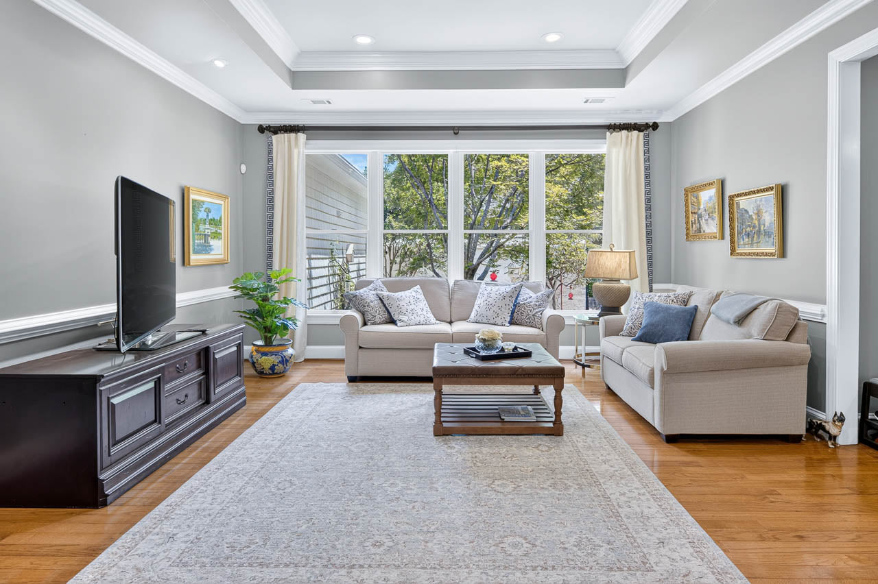 Bright living room with large windows, two beige sofas, a coffee table, a TV on a stand, framed artwork on the walls, a plant, and a light-colored rug on hardwood floors.