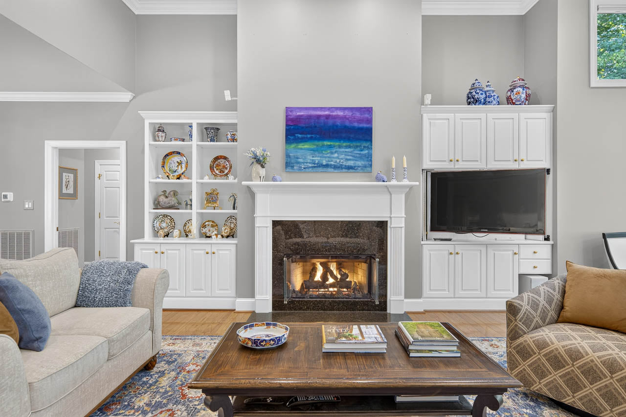 A cozy living room with a lit fireplace, white built-in shelves, a TV, a blue and purple painting above the mantel, a beige sofa, and a wooden coffee table with books and a bowl.