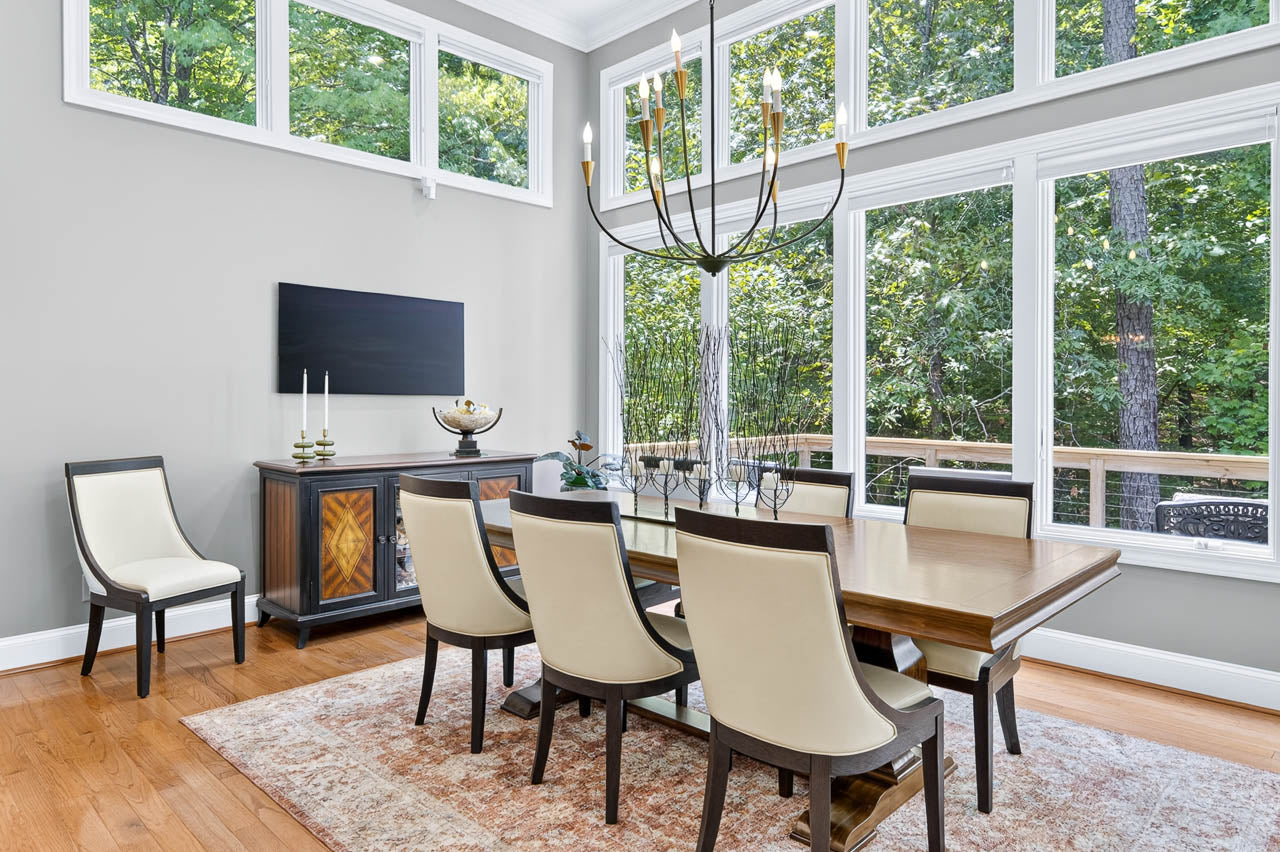 Modern dining room with a wooden table, eight cream-colored chairs, a large chandelier, rug, sideboard, candles, and large windows showing a green wooded view outside. Walls are light gray with hardwood flooring.