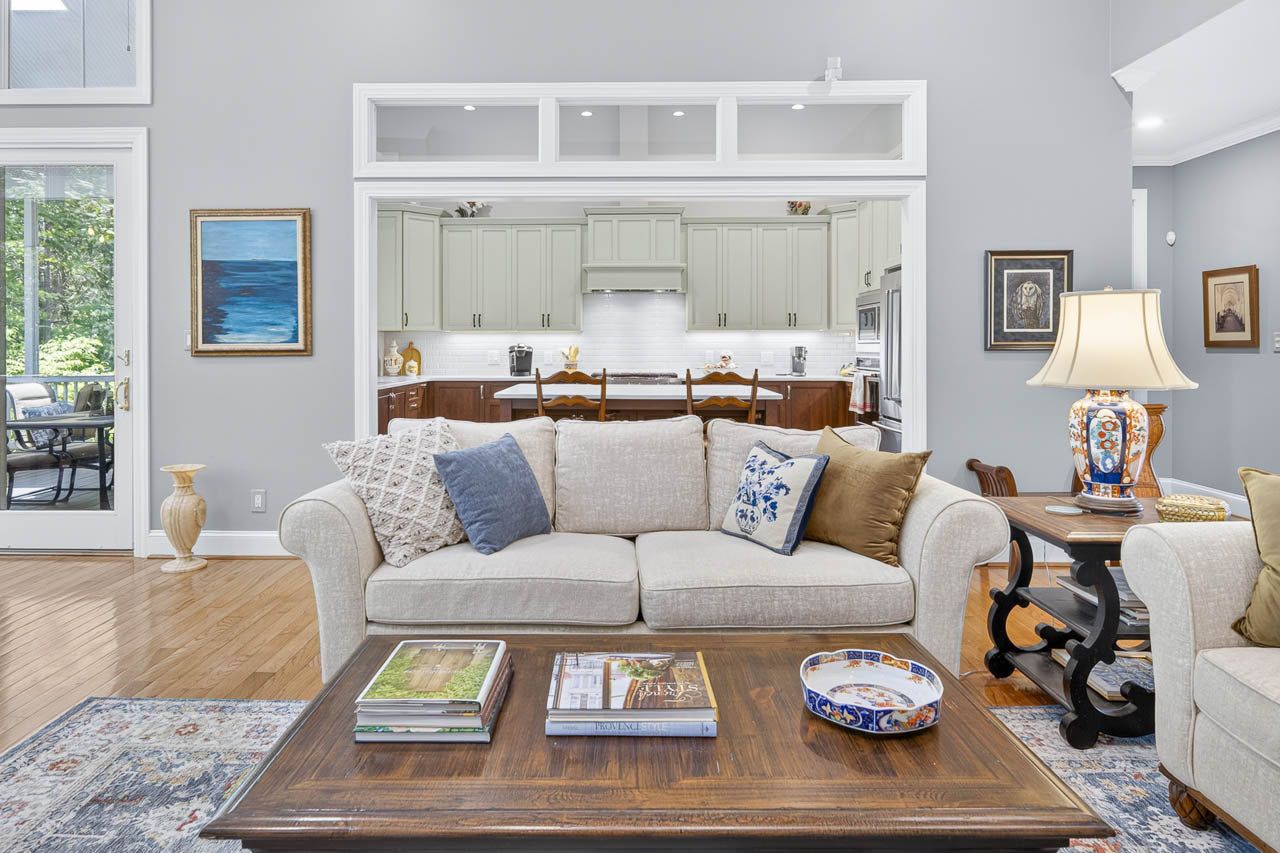 A cozy living room with a beige sofa, assorted pillows, a wooden coffee table with books and decor, and a view into a modern white kitchen. Art and lamps decorate the space.