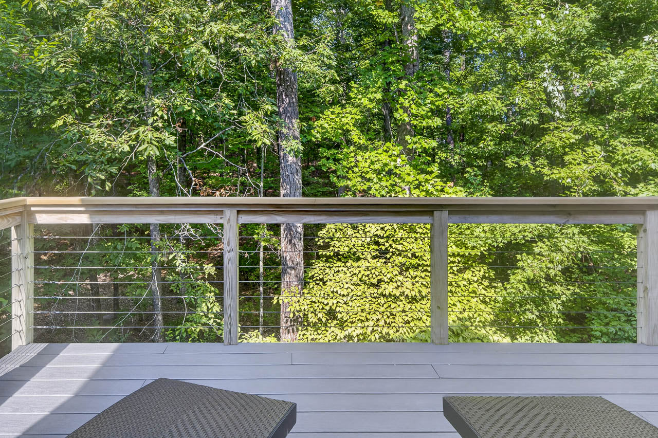 Wood deck with horizontal cable railing overlooks a lush, green forest with tall trees. Two woven lounge chairs are partially visible in the foreground. Bright, sunny day.