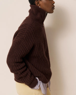 A person wearing the HARTFORD - MYPIO Sweater in Chocolate, styled over a striped shirt and paired with light-colored pants, stands in profile with hands in pockets against a neutral background.
