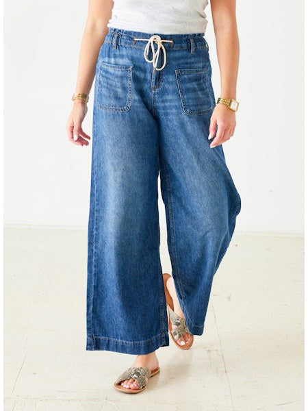 Wearing MERSEA&#39;s LONDONBERRY Drawstring Supersoft Denim Pant with wide legs and large front pockets, paired with a light grey top, gold jewelry, and brown sandals, a person poses against a plain light background.