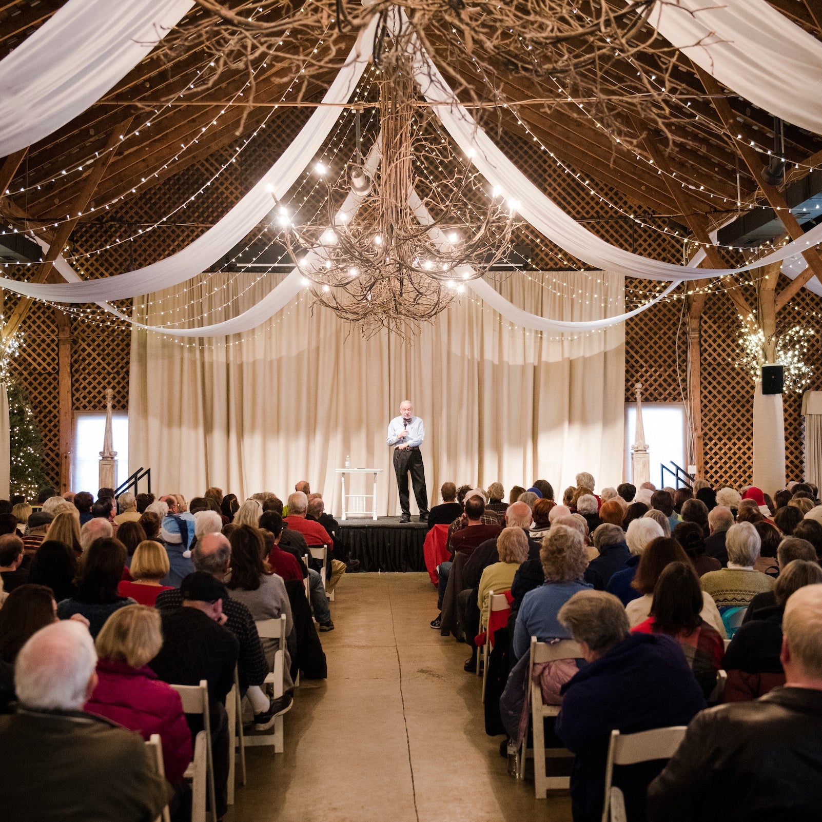 A man stands on a small stage speaking to a seated audience in a rustic hall decorated with draped white fabric, string lights, and a large antler chandelier.