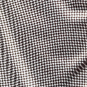 A man with short brown hair and a beard wears the COLLARS AND CO Semi Spread Collar Original Polo in Brown Houndstooth, standing in an urban setting with blurred buildings and warm outdoor lighting in the background.