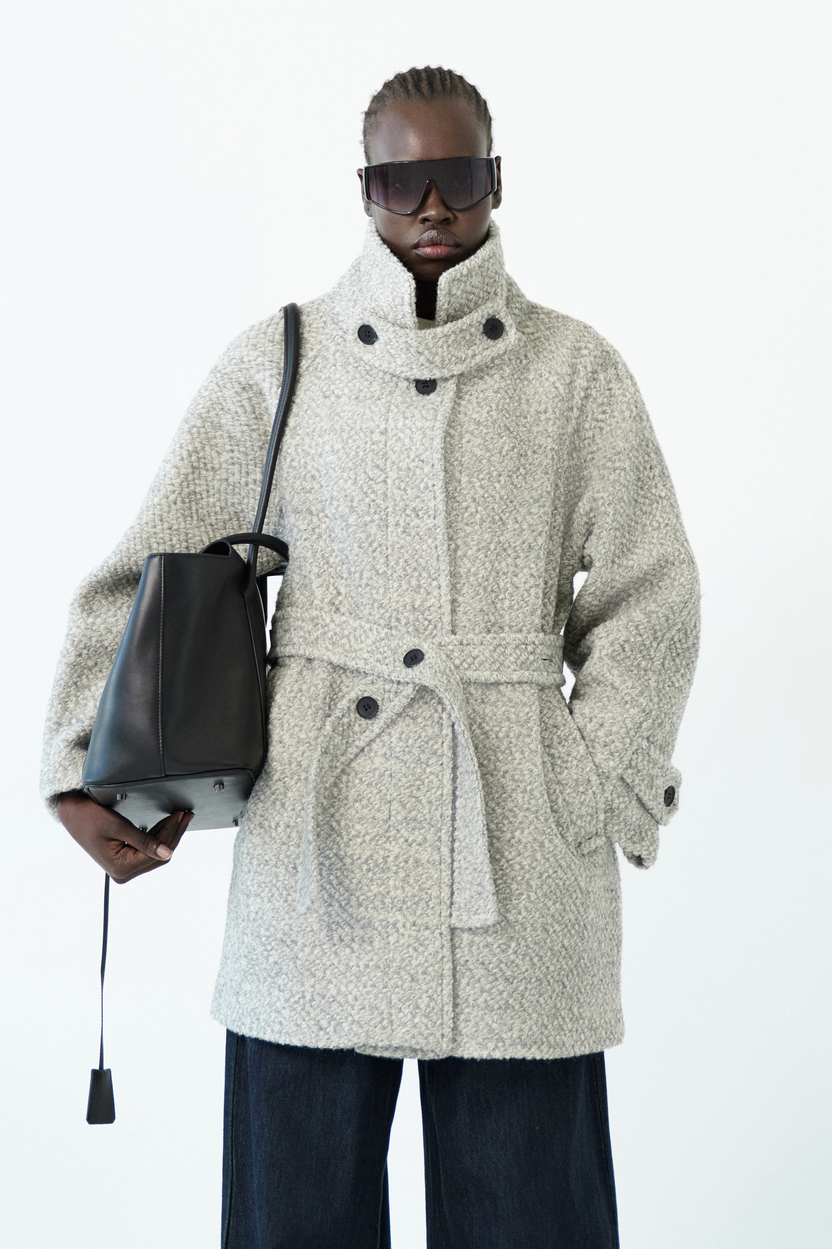 Close-up of the ELEH - THE AURELIA COAT in light gray wool blend, showing a fabric belt and black cuff button, hanging against a white wall.