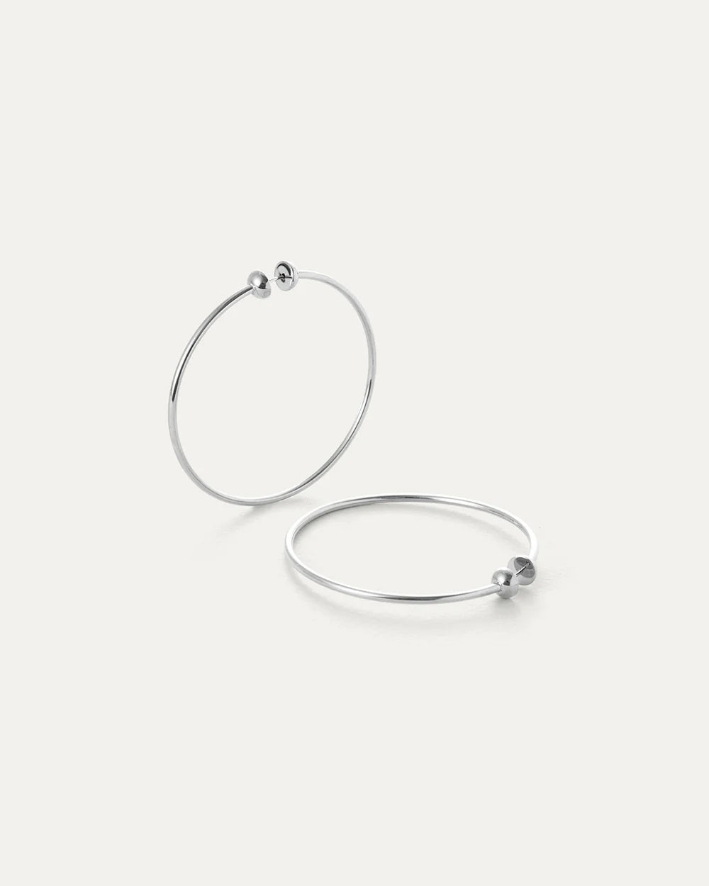 The JENNY BIRD - MEDIUM ICON HOOP EARRINGS, made from high polish stainless steel, feature two thin silver hoops with small beads at the clasp. Set against an off-white background, one earring is upright while the other lies flat.
