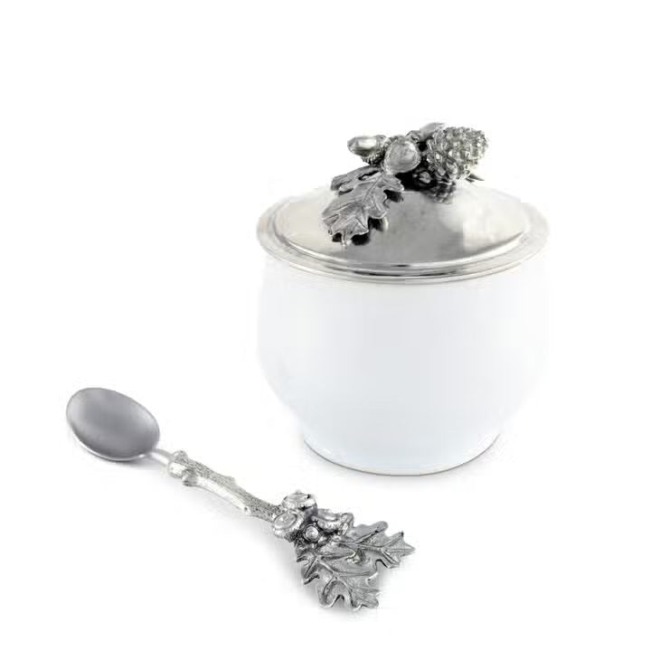The ACORN OAK LEAF SUGAR BOWL by VAGABOND HOUSE is a small fine porcelain bowl with a silver lid adorned with intricate decorative elements, including acorns and oak leaves. The lid&#39;s handle is an ornate oak leaf design, enhancing the bowl&#39;s elegant and detailed appearance, reminiscent of a luxury grade pewter Antler Creamer Set.