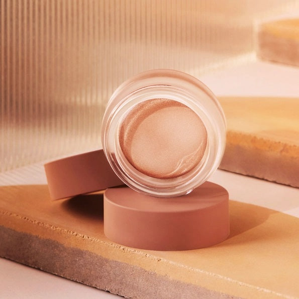 A small round container of MINORI - CREAM HIGHLIGHTER, CHAMPAGNE with a pink lid is shown partly open, revealing the shimmery champagne cream inside for a natural glow.