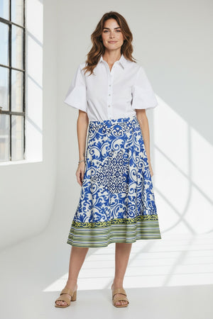 A person, whose head and feet are not visible, stands against a plain background wearing a white blouse and the HINSON WU - GLORIA SKIRT IN CERAMIC TILE PRINT midi skirt by HINSON WU, featuring a blue and white ceramic tile print with a green striped hem.