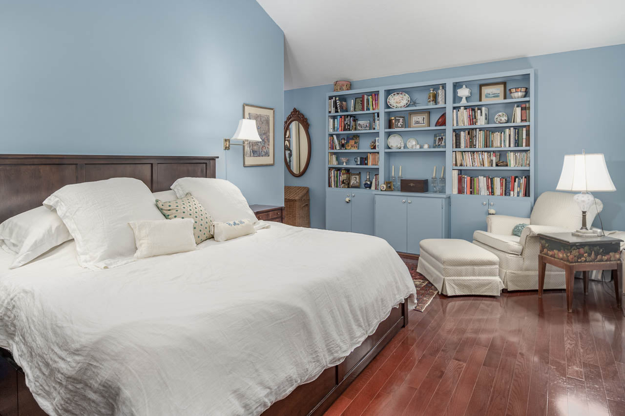 A cozy bedroom with light blue walls, a large bed with white bedding, wooden floors, built-in bookshelves filled with books and decor, and a white armchair with a matching ottoman by a lamp.