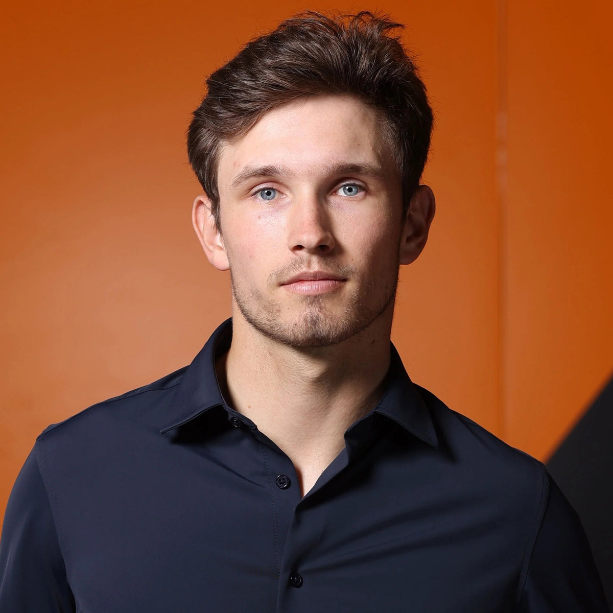 A man wearing the COLLARS AND CO - APEX PERFORMANCE DRESS SHIRT SEMI SPREAD IN NAVY stands in front of an orange race car holding a racing helmet, with an orange and black background behind him.