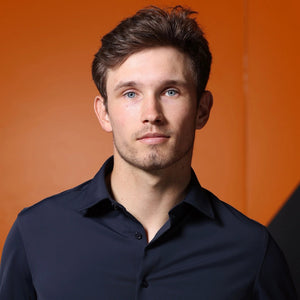 A man wearing the COLLARS AND CO - APEX PERFORMANCE DRESS SHIRT SEMI SPREAD IN NAVY stands in front of an orange race car holding a racing helmet, with an orange and black background behind him.