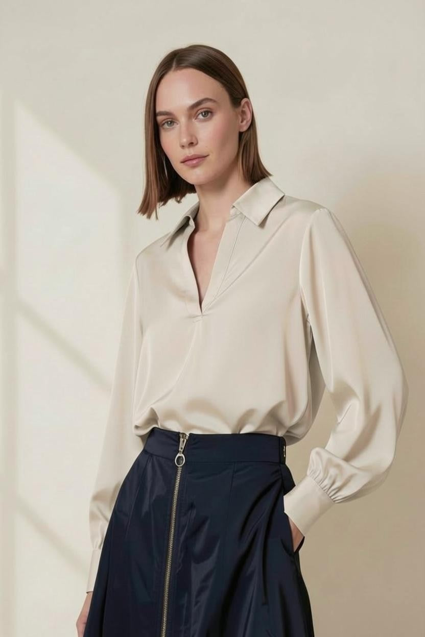 A woman with straight brown hair wears the VICARIO CINQUE Solei Taffeta Top in beige, featuring a collar and long sleeves, tucked into a high-waisted navy skirt with a front zipper. She stands against a light, neutral background.