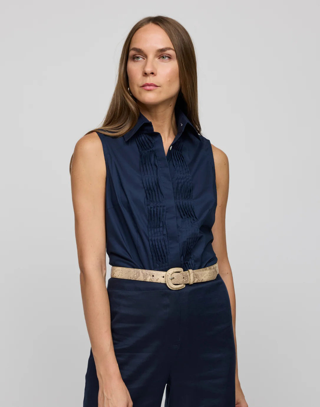 A woman with long hair wears the HINSON WU Lilith Sleeveless Shirt, made from stretch polished cotton with pleats and a beige belt, against a gray background. She exudes confidence as she gazes at the camera, highlighting the shirt&#39;s versatility.