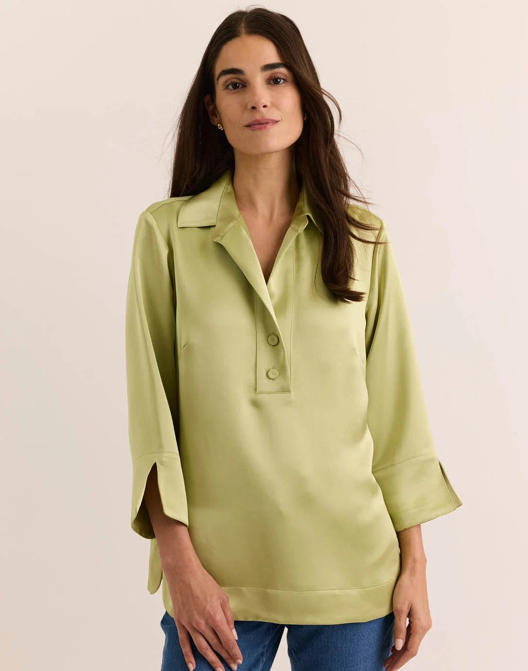 A woman with long brown hair is wearing the HINSON WU - AILEEN 3/4 Sleeve Liquid Satin Shirt in Pistachio, a light green collared blouse with three-quarter sleeves, paired with blue jeans as she stands against a plain, light-colored background. The shirt by HINSON WU features an elegant drape and luminous sheen, adding a touch of sophistication to her relaxed look.