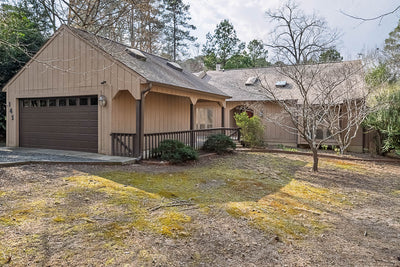 Single-story house with beige wood siding, a dark brown two-car garage, a ramp leading to the front door, and a leafless tree in the front yard. The home is surrounded by trees and has a shaded, earthy front lawn.