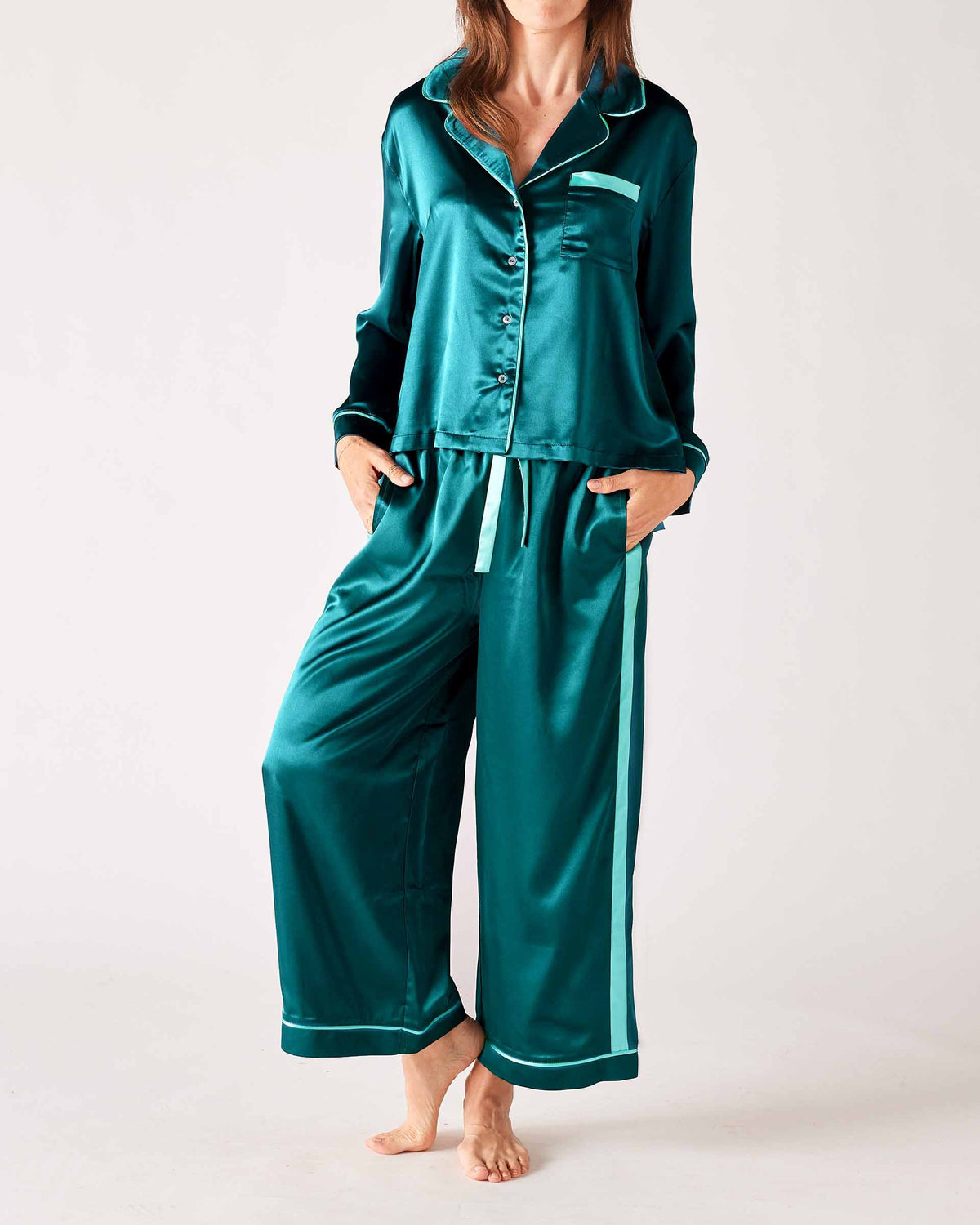 A woman models the MERSEA - OVER THE MOON SATIN SAILOR PAJAMA SET in teal with white trim, featuring a long-sleeved button-up top and elastic waist bottoms. She stands barefoot with her hands in the pockets, highlighting the silky smooth fabric.