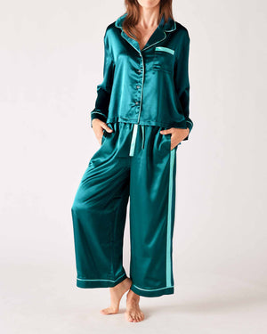 A woman models the MERSEA - OVER THE MOON SATIN SAILOR PAJAMA SET in teal with white trim, featuring a long-sleeved button-up top and elastic waist bottoms. She stands barefoot with her hands in the pockets, highlighting the silky smooth fabric.