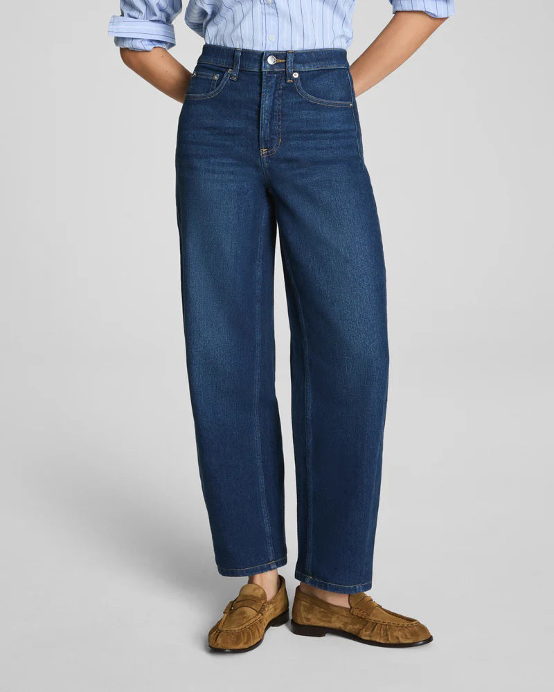 A person wearing SPANX BARREL LEG JEANS by SPANX in dark blue, paired with a light blue rolled-up sleeve shirt and brown moccasin loafers, shown from the back.