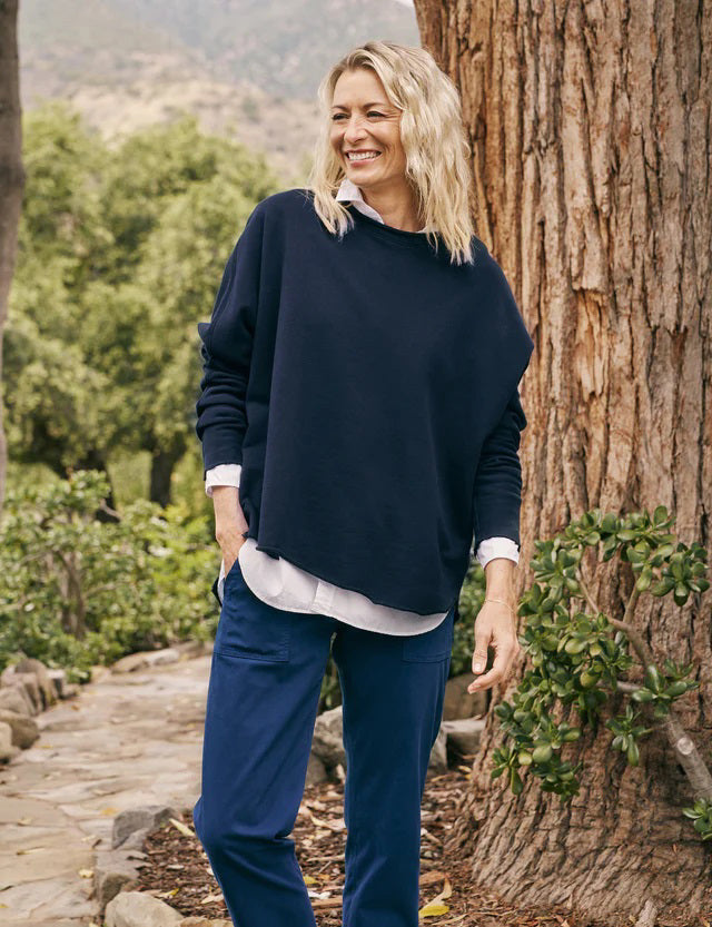 A person with long, wavy blonde hair is standing outdoors near a large tree, wearing the FRANK &amp; EILEEN ANNA LONG-SLEEVE CAPELET in navy over a white shirt and blue pants. The background showcases green foliage and a stone path. The person is smiling and gazing to the side.