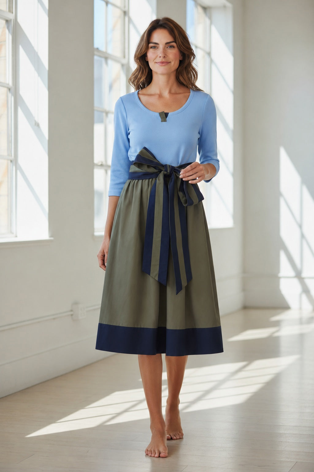 A woman with wavy brown hair stands barefoot in a sunlit room with large windows, wearing the ROSSO35 Color Block Dress in Light Blue Military Green by ROSSO 35 SILKY. This effortlessly elegant midi dress features an olive green and light blue color-block design accented by a large navy bow—making it a perfect choice for special occasions.