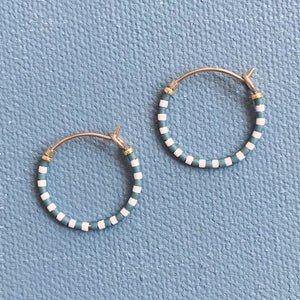 The ALICE RISE LITTLE COLORLOOP EARRINGS IN SLATE BLUE by ALICE RISE, featuring gold hoops with alternating white and teal Japanese glass beads, are displayed on a textured light blue surface, highlighting their handmade elegance.