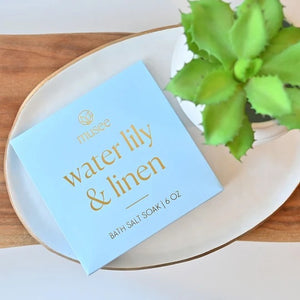 A blue box labeled “MUSEE - WATER LILY & LINEN MINI BATH SALT SOAK” by MUSEE sits neatly, with a small heap of bath salts—a calming blend of natural ingredients—spilled in front, inviting relaxation.