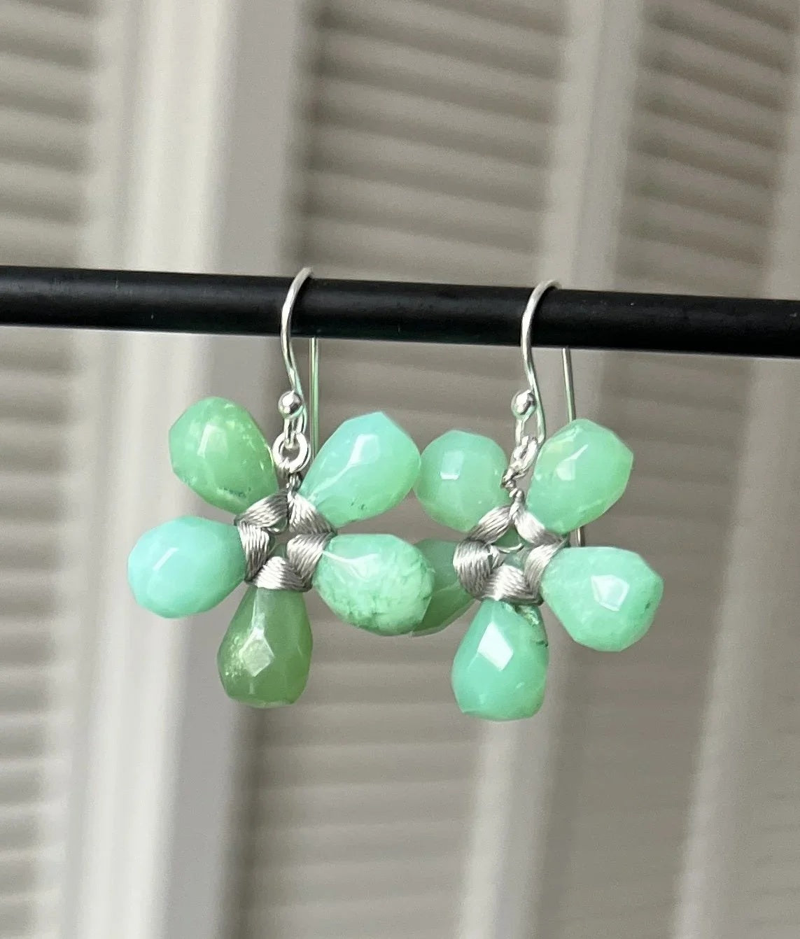 The ANN LIGHTFOOT - FACETED CHRYSOPRASE FLOWER EARRINGS by ANN LIGHTFOOT feature blossom-shaped designs crafted from five faceted green chrysoprase stones with delicate silver wire wrapping at the center, elegantly hanging from sterling silver hooks against a softly blurred, neutral background.
