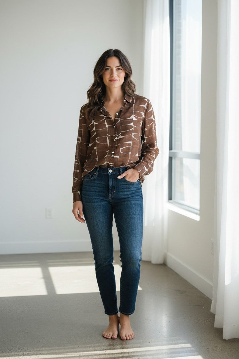 A woman stands barefoot indoors, wearing the HARTFORD - CINEMA SHIRT IN BROWN and blue jeans. She has wavy dark hair and is smiling slightly, with natural light coming through large windows and white curtains.