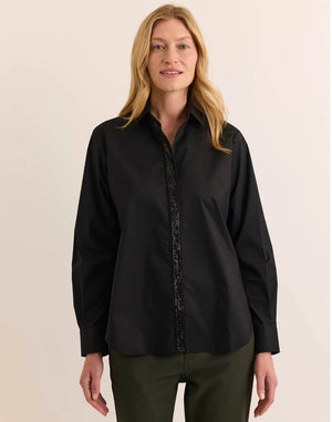A woman wearing a black HINSON WU - ESTHER LONG SLEEVE SHIRT WITH SEQUIN TRIM under a black velvet blazer, paired with olive green trousers, black high-heeled sandals, and holding a black clutch.