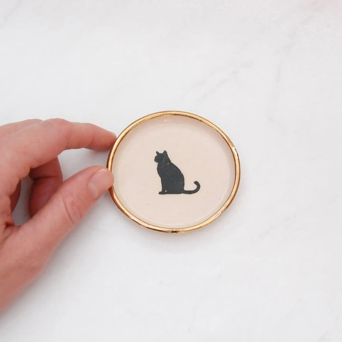 The COMPOST DESIGN Sitting Cat Tiny Dish 3&quot; features a black silhouette of a sitting cat on a cream background, framed by an elegant 22k gold edge. Perfect for decorative use, it rests beautifully on light, textured surfaces.