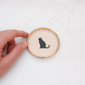 The COMPOST DESIGN Sitting Cat Tiny Dish 3" features a black silhouette of a sitting cat on a cream background, framed by an elegant 22k gold edge. Perfect for decorative use, it rests beautifully on light, textured surfaces.