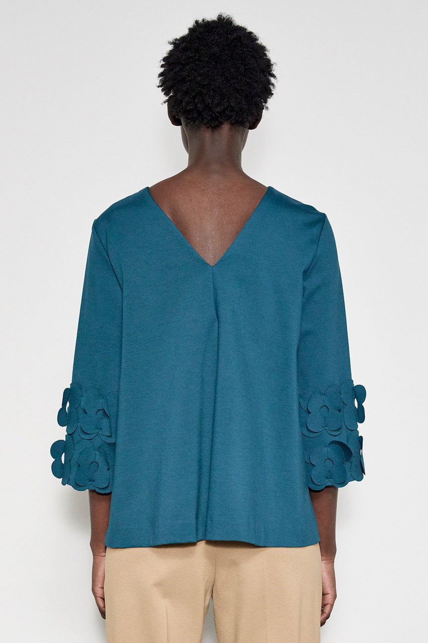 A person with short curly hair stands in profile against a white background. They are wearing the MEIMEIJ - 3/4 Sleeve Back to Front Top with Lasercut Flower Detail, a loose-fitting teal top featuring flower embellishments on the sleeves, paired with light beige pants.