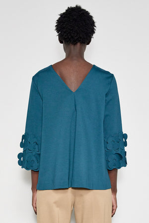 A person with short curly hair stands in profile against a white background. They are wearing the MEIMEIJ - 3/4 Sleeve Back to Front Top with Lasercut Flower Detail, a loose-fitting teal top featuring flower embellishments on the sleeves, paired with light beige pants.