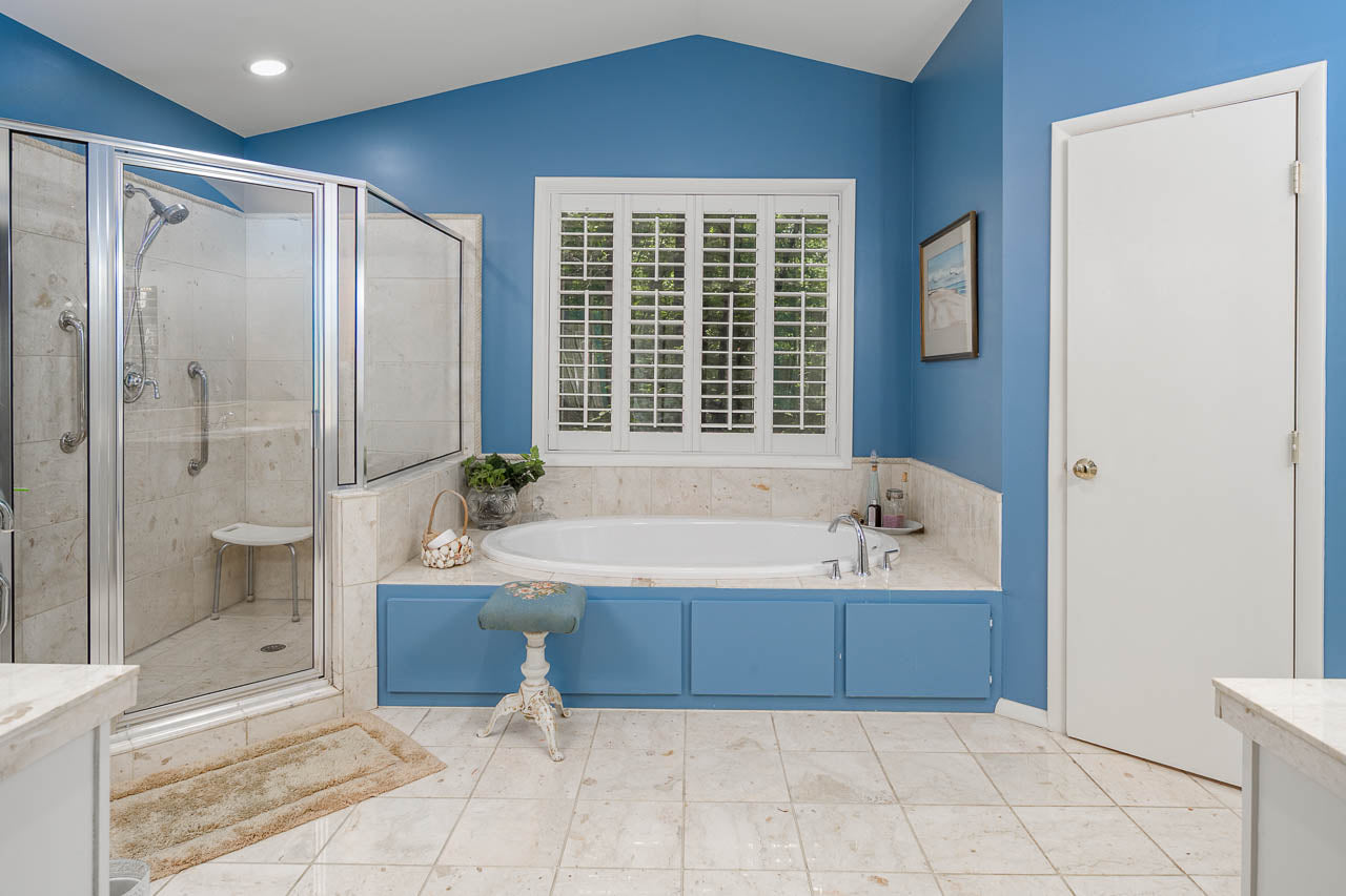 A spacious bathroom with blue walls, a large glass shower, a bathtub with marble surround, white tile floor, a small stool, and a window with white shutters.