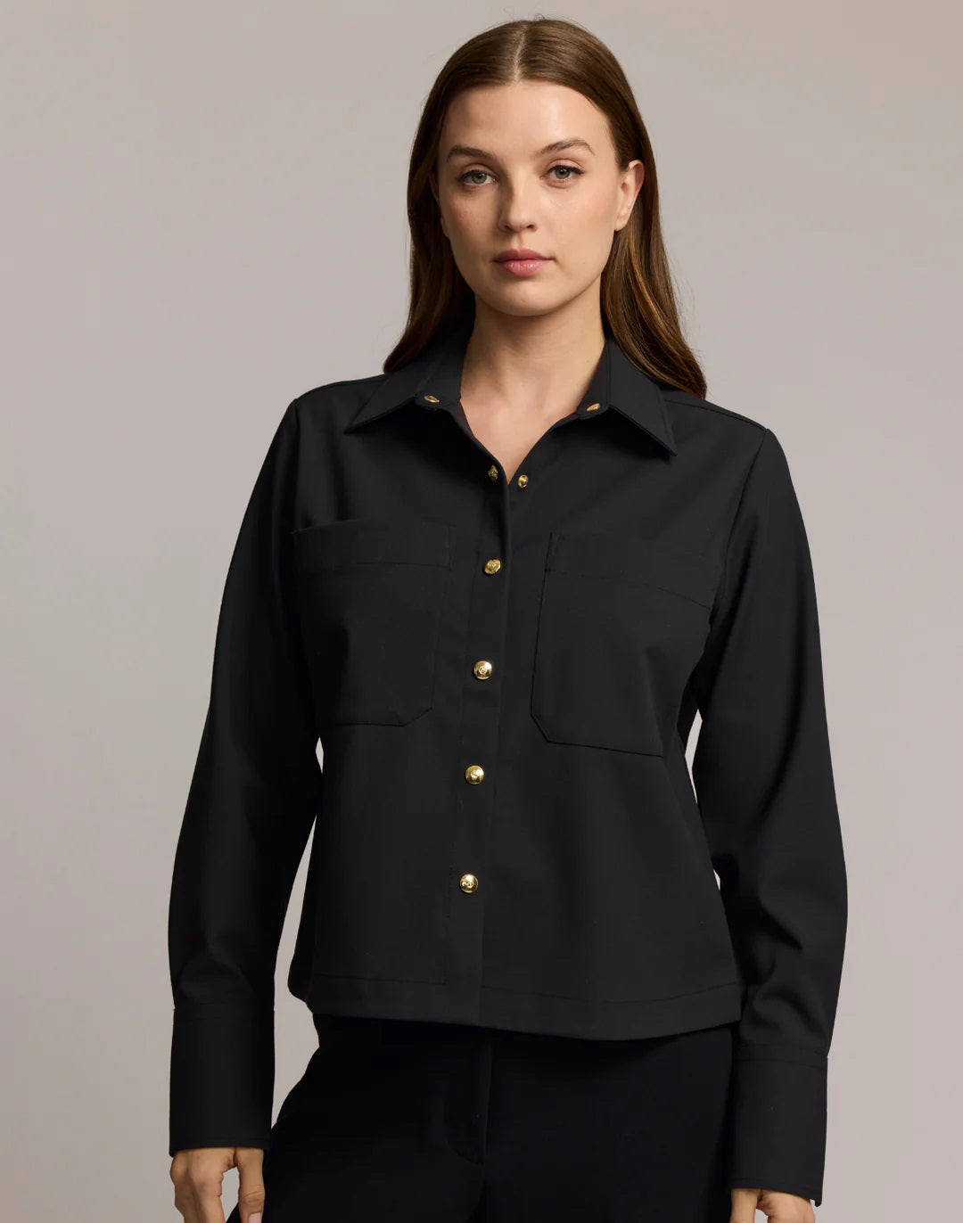 A woman with long brown hair models the HINSON WU - ASTRID LONG SLEEVE TRAVELER&#39;S TWILL SHIRT JACKET in dark green over an olive top and blue jeans, showcasing versatile HINSON WU travel style with gold hoop earrings against a neutral background.
