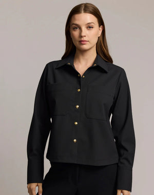 A woman with long brown hair models the HINSON WU - ASTRID LONG SLEEVE TRAVELER'S TWILL SHIRT JACKET in dark green over an olive top and blue jeans, showcasing versatile HINSON WU travel style with gold hoop earrings against a neutral background.
