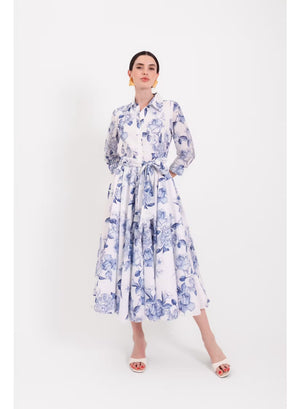 Model wearing the LAVI - SPRITZ DRESS IN BLUE FLORAL by LAVI—a white, long-sleeve, button-down midi shirt dress featuring blue botanical floral prints and a waist tie—paired with white heeled sandals and gold statement earrings, standing against a plain white background.