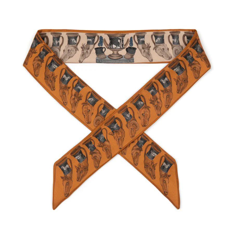 A person is wearing a white short-sleeved top paired with a pleated white skirt. They have SABINA SAVAGE ENGLAND's THE TEMPLE OF PEGASUS SILK RIBBON scarf, which features an orange and gray pattern reminiscent of ancient Greek art, tied neatly around their neck. The background is plain and neutral-colored.