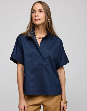 A woman with long brown hair is seen wearing the HINSON WU Cara Elbow Sleeve Shirt in Stretch Polished Cotton Navy—a loose-fitting, oversized short-sleeve shirt—paired with tan pants and gold jewelry, standing against a plain light gray background.