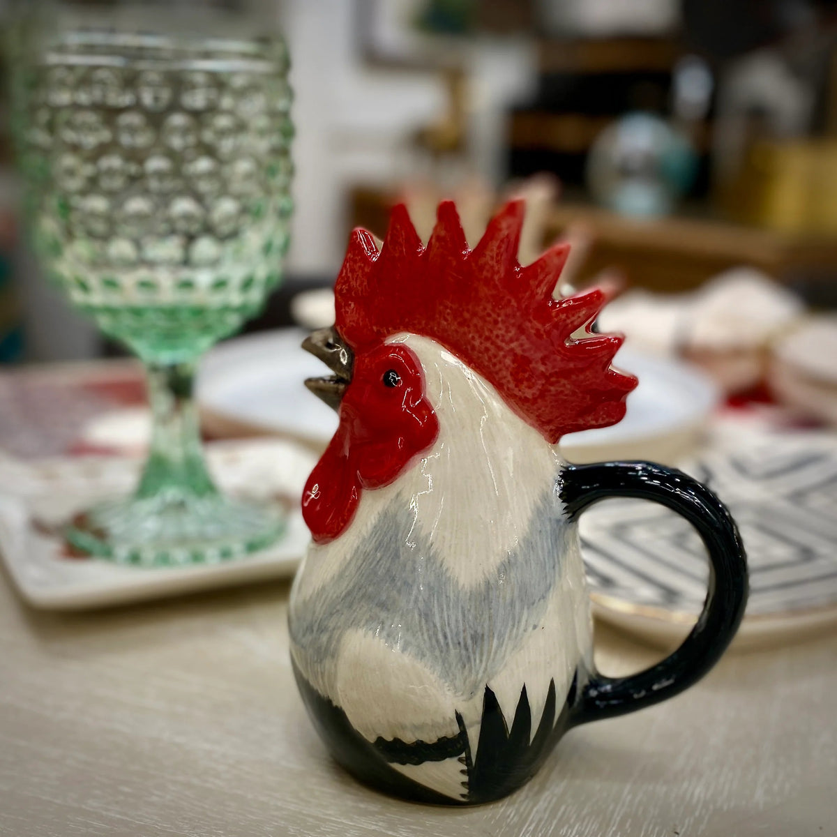 The QUAIL CERAMICS - DORKING ROOSTER JUG by QUAIL DESIGNS, a hand-painted stoneware jug with a red comb and black handle, sits on the table beside a green glass and patterned plates—a charming British tea accessory.